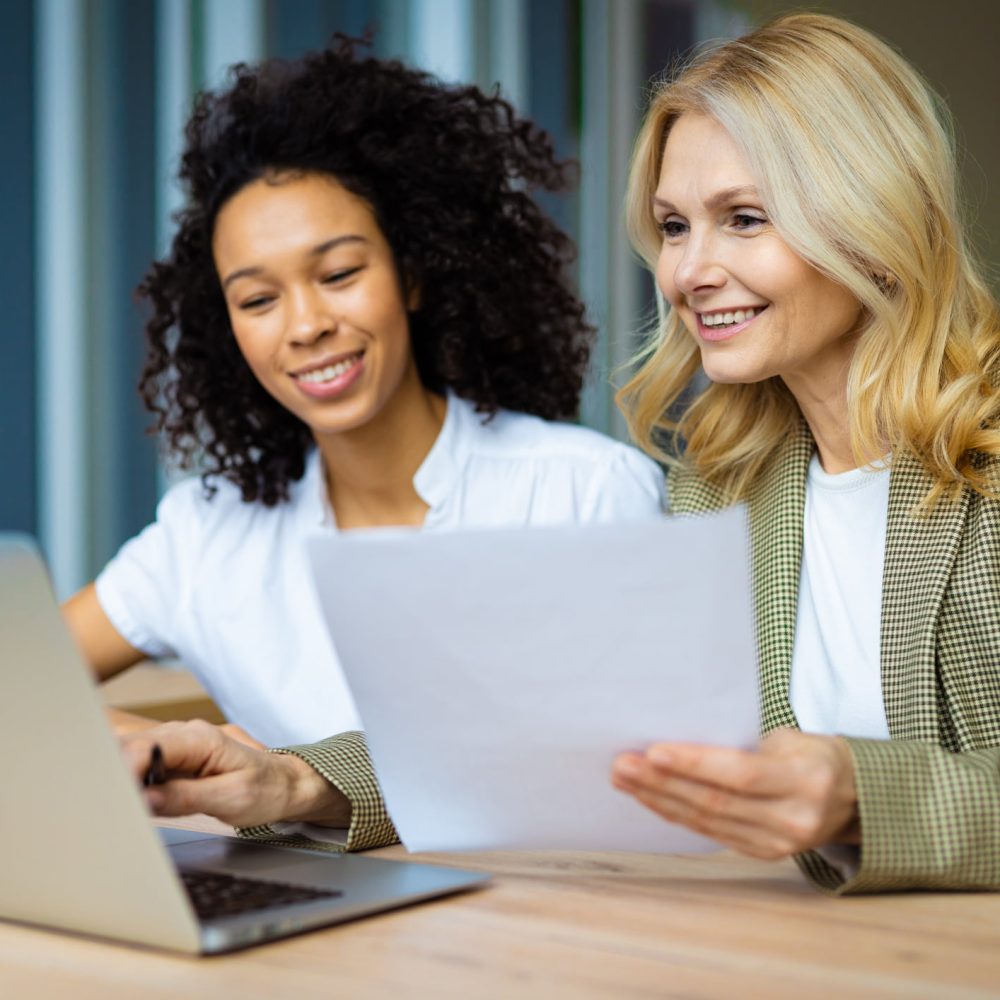 Beautiful adult businesswomen with elegant dress sitting at computer desk in the office - Senior caucasian woman and younger african american female working at laptop online, concepts about business and technology